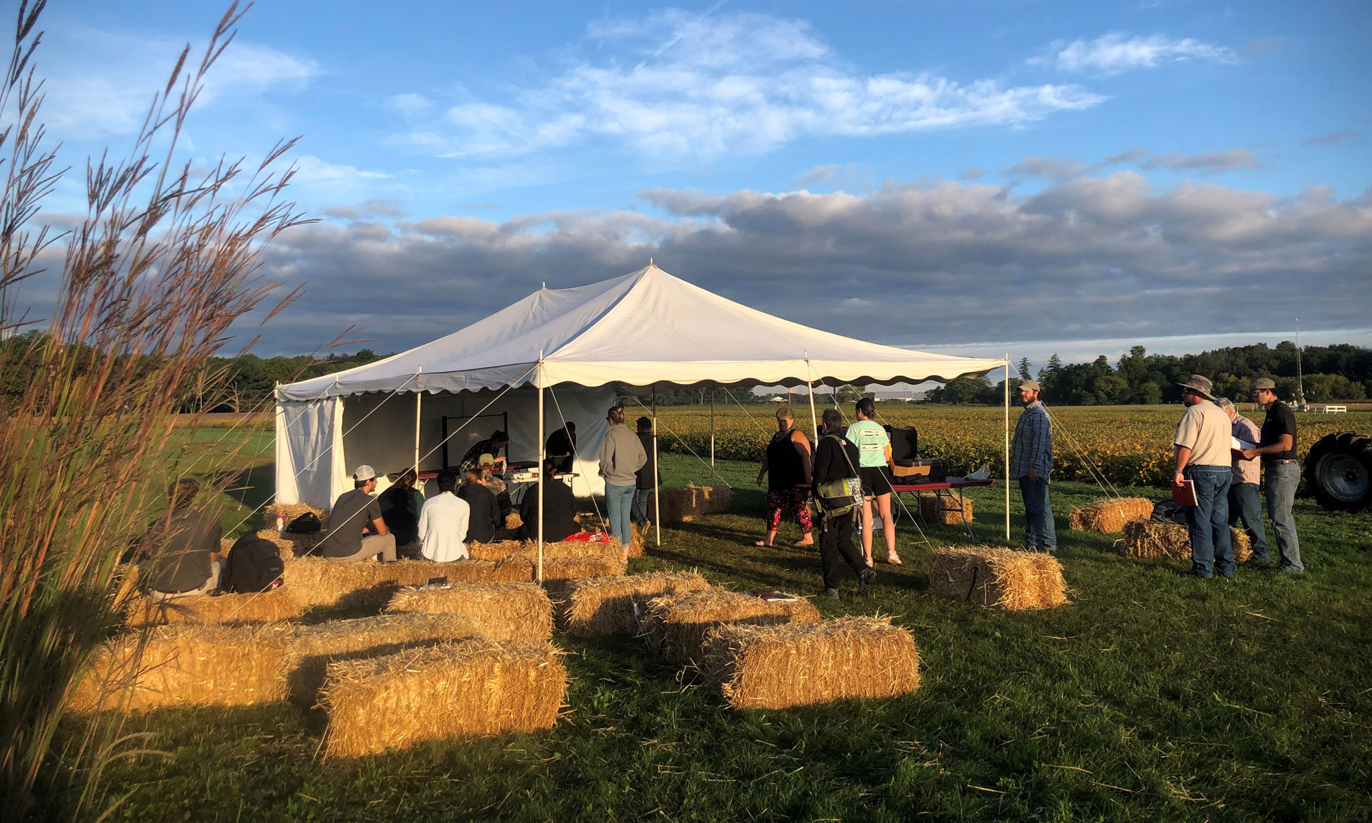 People inside and outside of a tent setup in a field with bales of straw to sit on.