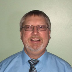 A man with short hair glasses and a beard wearing a light blue dress shirt and a tie