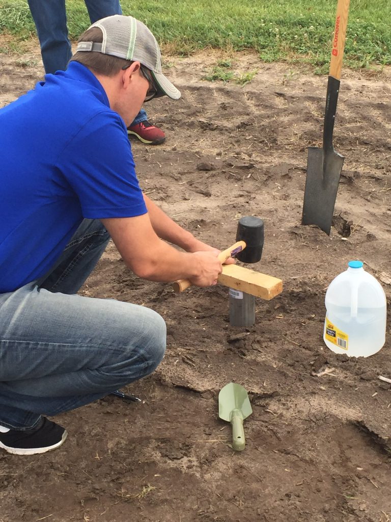 A man squatting down in a field using a rubber mallet and block of word to do a water infiltration test.